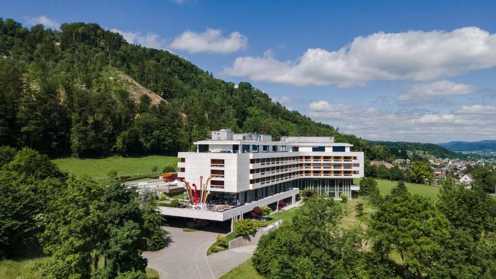 Prédio branco do FIVE Zurich, um dos hotéis em Zurique, em um dia ensolarado, com bastante natureza verde em volta e vista no horizonte das casas da cidade