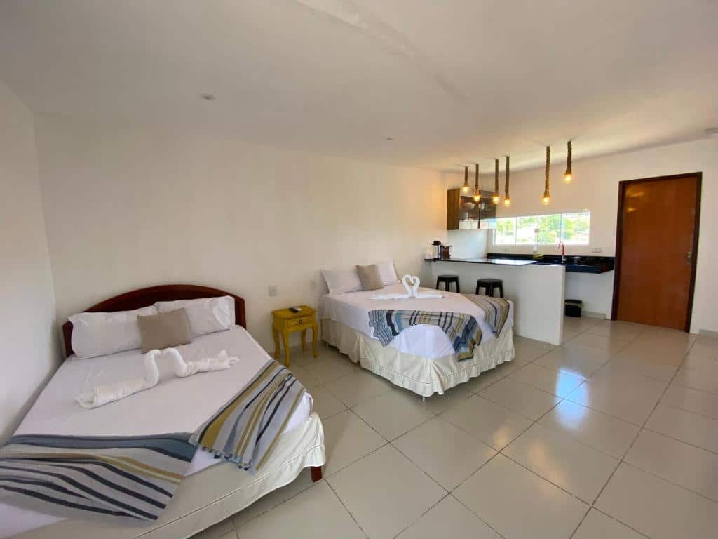 Quarto do Flat Por do Sol com uma cama de casal no canto esquerdo, encostada na parede e uma cama de solteira do lado direito. No meio delas há uma mesinha. Ao lado da cama de solteiro existem dois banquinhos e um balcão que separa a área da cozinha, que possui também uma pia grudada na parede do fundo. Ao lado da pia está a porta. Essa é uma foto para ilustrar o post de pousadas em Baía Formosa, RN.