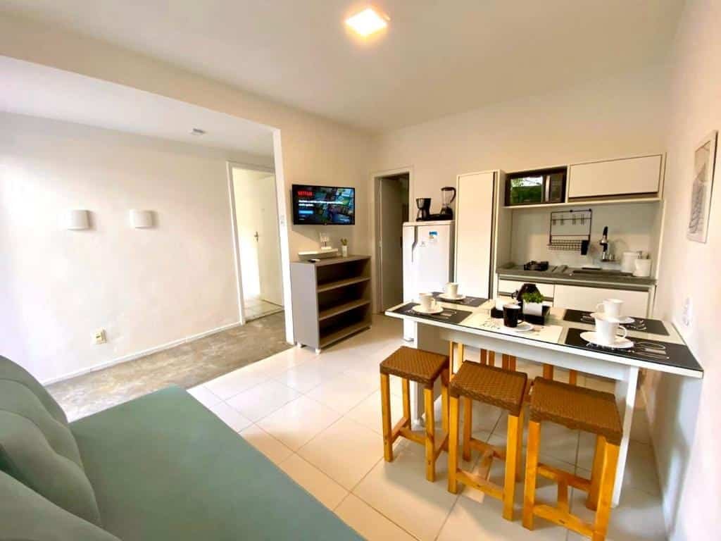 Cozinha do Flats Morro de São Paulo. No fundo, no lado direito a pia com utensílios da cozinha. No meio uma mesa posta com banquetas. Do lado esquerdo uma cômoda com uma televisão e o corredor do apartamento.