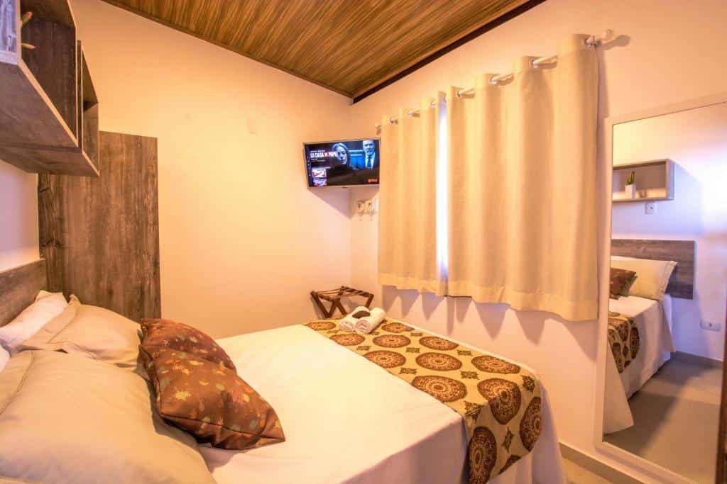 Quarto do Flats Morro de São Paulo. Uma cama de casal do lado esquerdo, prateleiras em cima. De frente um espelho, uma janela com cortinas fechadas e uma televisão.