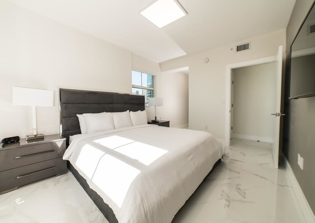 A imagem mostra um quarto do Fortune House Hotel com uma cama de casal grande, coberta com roupa de cama branca. A cabeceira da cama é acolchoada em um tom escuro, contrastando com as paredes claras. Há duas mesas de cabeceira pretas com lâmpadas brancas modernas. O piso é de mármore branco com veios cinza, dando um toque sofisticado ao ambiente. À direita, há uma porta aberta que leva a outra parte do quarto. A luz natural entra por uma janela, iluminando o espaço de maneira suave e acolhedora.
