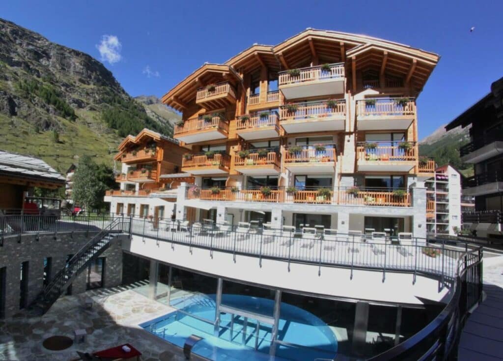 Imagem da frente do Alpenhotel Fleurs de Zermatt durante o dia com piscina localizada abaixo da propriedade e acima os apartamentos.