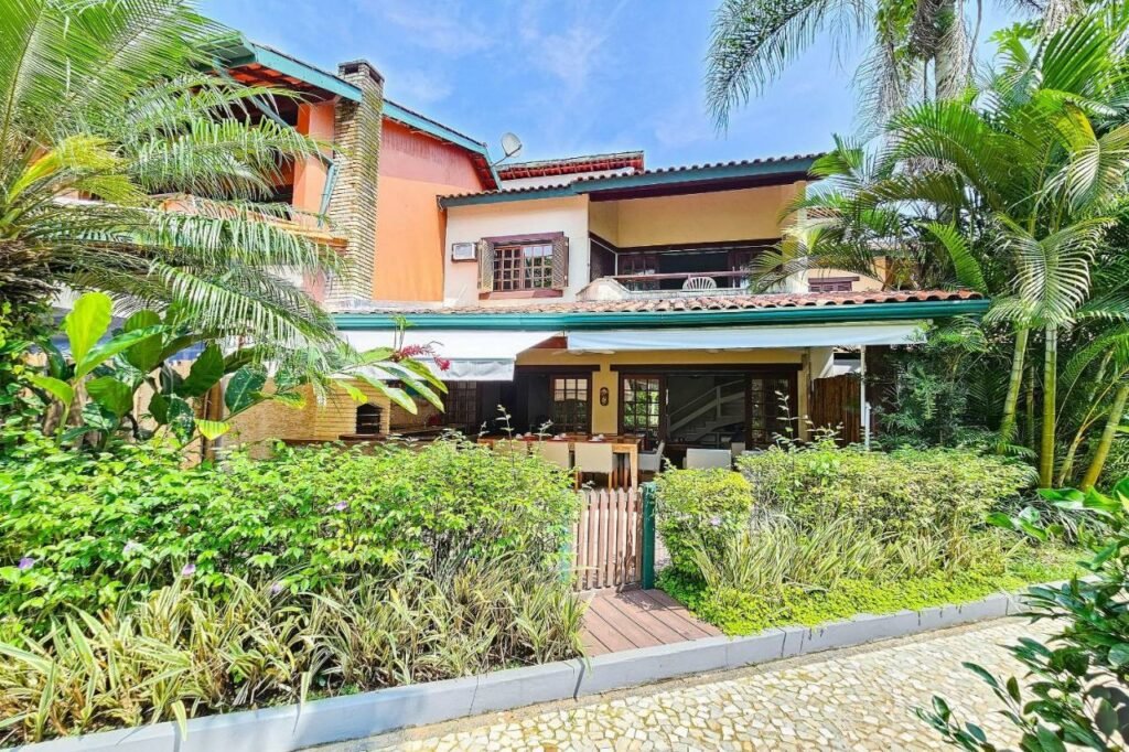 Frente da Casa com varanda e churrasqueira em São Sebastião durante o dia com um portão de madeira a frente em volta de um jardim e ao fundo a casa.