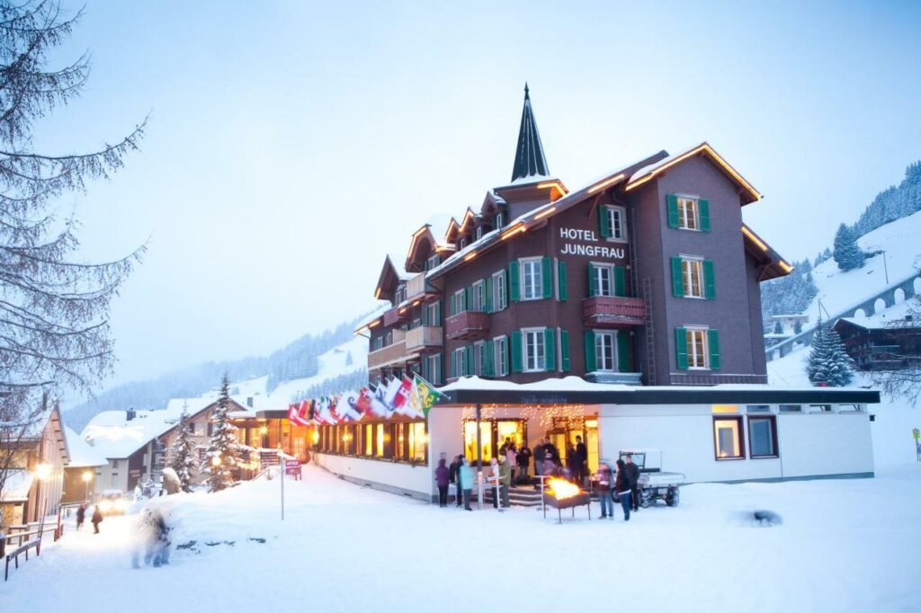 Imagem da frente do Hotel Jungfrau Mürren no final do dia com neve a frente e algumas pessoas e ao fundo a estadia. Representa hotéis em Lauterbrunnen.