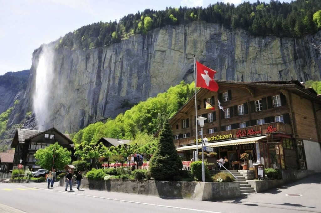 Imagem da frente do Hotel Schützen Lauterbrunnen com a estadia do lado direito da imagem ao fundo do hotel uma parede rochosa.