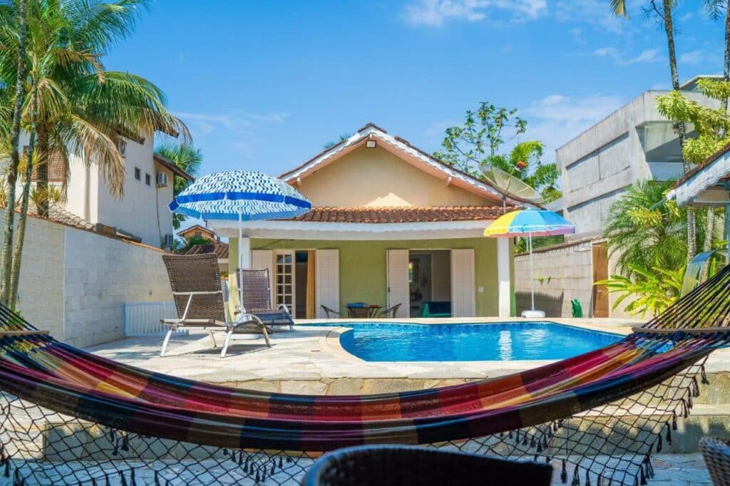Area de lazer da Maravilhosa casa no Cond. fechado Morada da Praia durante o dia com rede a frente, ao fundo do lado esquerdo da imagem duas cadeiras com guarda-sol e do lado direito a piscina ao fundo a estadia.