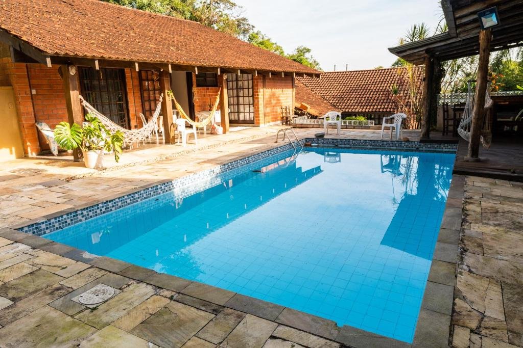 vista da piscina na pousada Gajah em Joinville com uma varanda coberta à esquerda, mostrando algumas redes penduradas nas colunas de sustento do telhado. O sol está batendo bem na entrada da varanda e numa parte da piscina retangular, em frente a varanda.