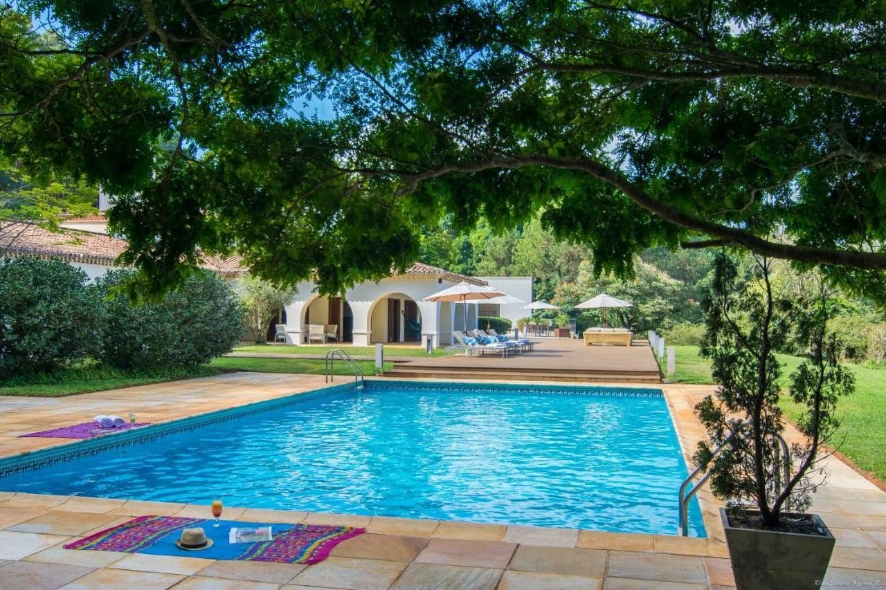Uma piscina no quintal do Garden Hill Hotel e Golfe, um dos hotéis fazenda em Minas Gerais, com espreguiçadeiras, guarda-sóis e duas toalhas no deck da piscina. Uma casa e árvores são visíveis ao fundo.