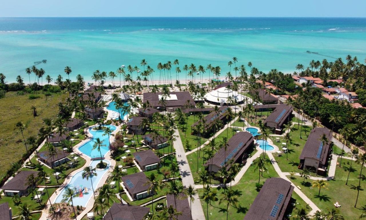 A imagem mostra uma vista aérea do Grand Oca Maragogi Resort, situado ao lado de uma praia de águas turquesas. O resort é composto por várias estruturas com telhados de telha escura, dispostas simetricamente ao redor de várias piscinas azul-claras. As piscinas são conectadas por caminhos pavimentados ladeados por palmeiras altas e gramados bem cuidados. Além das piscinas, a área é cercada por uma vegetação densa e uma praia de areia branca que se estende ao longo do horizonte. O cenário é paradisíaco, com um contraste vibrante entre o azul do mar, o verde da vegetação e as cores vivas do resort.