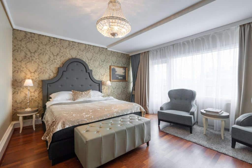 Quarto do Grand Hotel com acomodações sofisticadas, cama de casal, duas cômodas ao lado com luminária, duas poltronas cinzas do lado direto perto da janela.