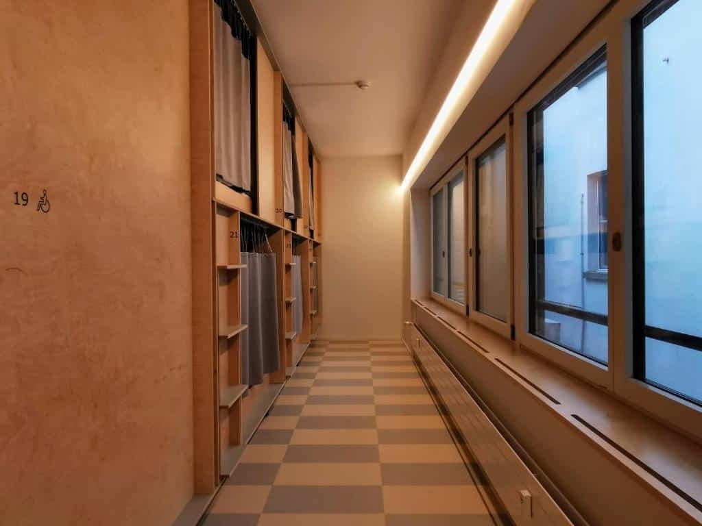 Corredor iluminado do Green Marmot Capsule Hotel, em Zurique, sendo de um lado as cápsulas para dormir com cortinas cinzas e do outro janelas de vidro com vista para um prédio branco