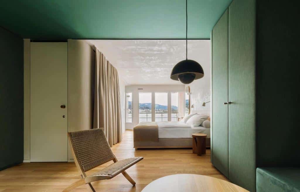Foto do Hermitage Lake Lucerne, para representar o post de hotéis em Lucerna. Em primeiro plano, na esquerda, vemos a porta do quarto. Na esquerda está o guarda-roupa, esse que é verde escuro. No canto inferior direito há uma mesa de madeira e uma cadeira. Já no fundo do quarto, após virar para a direita depois de adentrar a porta, há uma cama box de casal e varanda com vista para o lago.