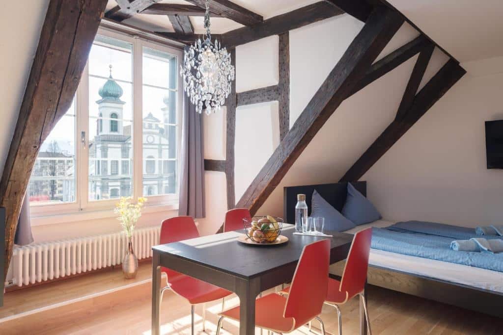 Um estúdio do HITrental Chapel Bridge Apartments. Na esquerda há uma grande janela para a cidade de Lucerna, e que permite entrar bastante luz natural. Na sua frente há uma mesa de madeira com quatro cadeiras. Em cima dessa mesa há uma cesta com fruta e um garrafa d