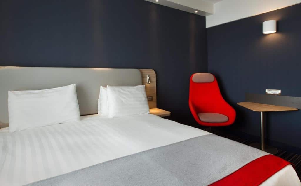 Foto de um quarto no Holiday Inn Express Geneva Airport, para ilustrar o post de hotéis em Genebra. A cama é box de casal, e vemos ela de frente, ocupando a maior parte esquerda da foto. Na direita, há uma poltrona vermelha com mesa. As paredes do quarto são azul escuro.