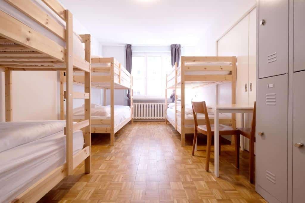 Foto do Barabas Luzern. Há três beliches no quarto e uma janela ano centro delas. Também há uma mesa com duas cadeiras na direita, na frente de uma das beliches. E do lado dessa mesa há um armário com cadeado de metal.