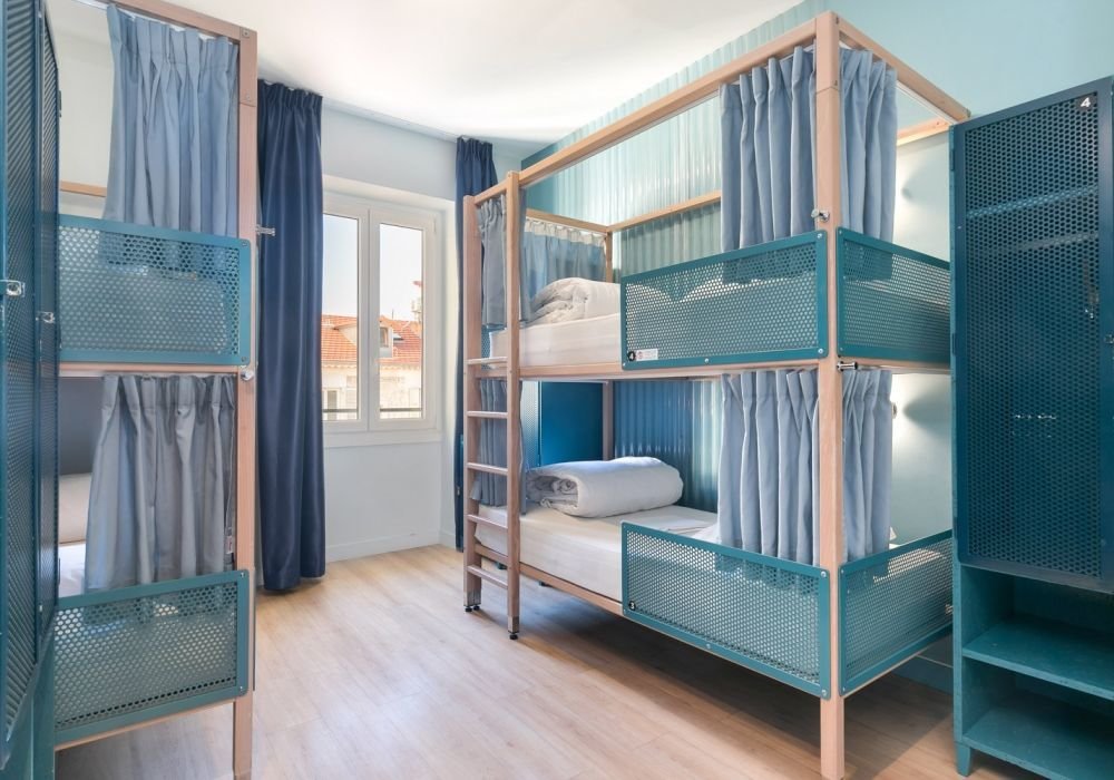 Imagem de um dos quartos do Hostel Ozz Nice. No lado direito há uma beliche com um armário ao lado, no fundo há uma janela com longas cortinas e no lado esquerdo há mais uma beliche com um armário ao lado.