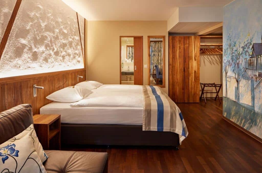 Suíte Júnior do Hotel Adler Zürich, de 28 m², com chão de madeira, cama de casal, sofá, espelho, banheiro e uma parede com pintura azulada de uma casa com árvores na frente