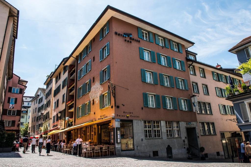 Esquina onde fica o prédio (de quatro andares) avermelhado do Hotel Adler Zürich, em Zurique, num dia ensolarado, com algumas pessoas andando e mesas ao lado do andar térreo