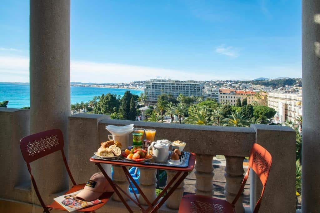 Varanda do Albert 1er. Há uma mesa com pães, frutas, suco, iogurte, café e ovos. Também há duas cadeiras na varanda e uma vista para o mar.