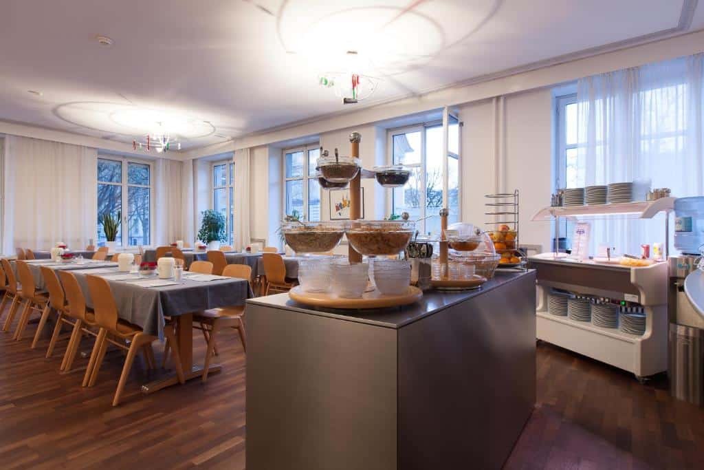 Imagem do restaurante do Hotel Alpha, e o buffet de café da manhã presente nele. O balcão é de metal e possui tigelas com várias opções de comida. Atrás, vemos as mesas e cadeiras. Também possui várias janelas espalhadas pelo ambiente.