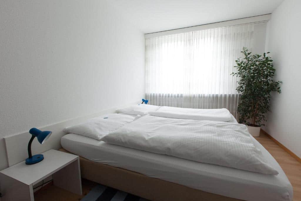 Foto do Hotel Alpha, para representar o post de hotéis em Lucerna. A cama box de casal está encostada na parede na esquerda, e há uma mesa de cabeceira com abajur em cima em seus dois lados. Na direita, no fundo da foto, há uma janela com cortinas brancas e um vaso de planta encostado no canto direito.