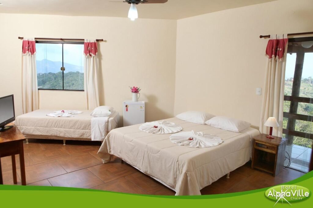 Um quarto do Hotel Alpha Ville Chalés com duas camas, uma TV em uma mesa de madeira, uma mini-geladeira e uma janela com vista para a montanha. Toalhas dobradas nas camas.