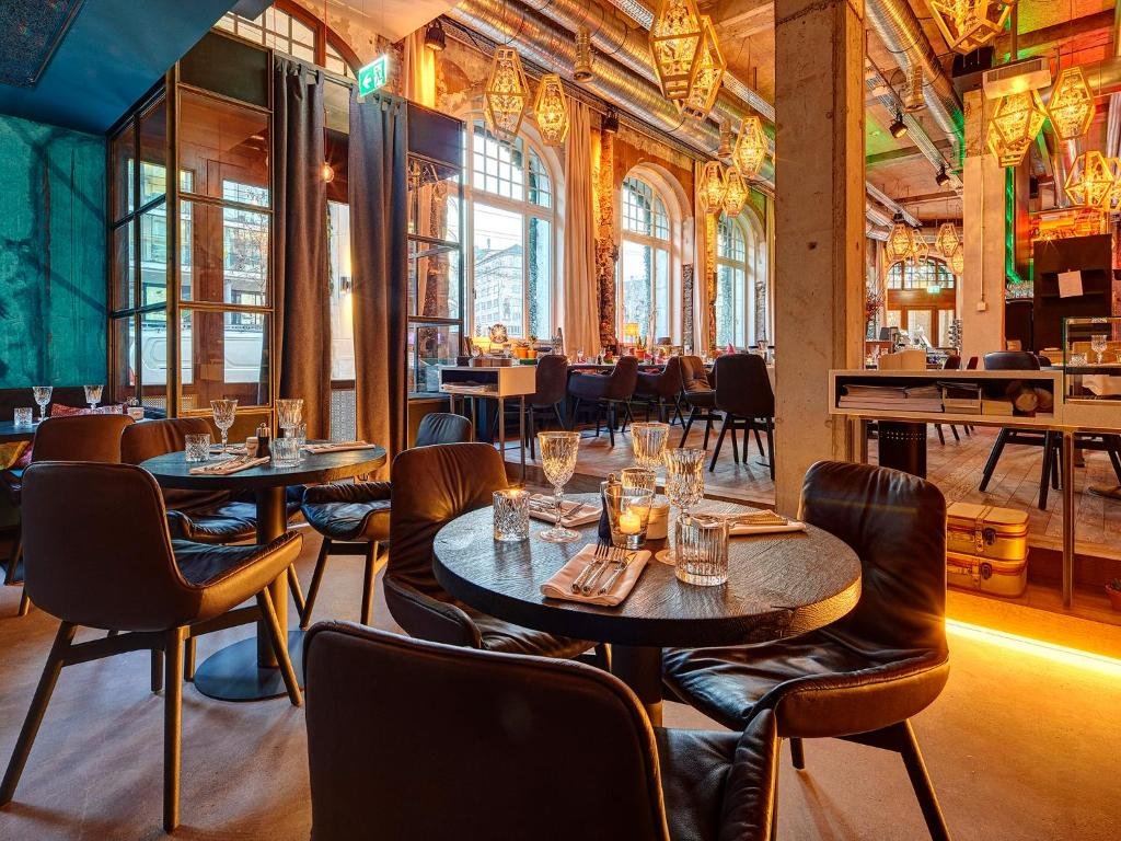 Um restaurante sofisticado no Hotel Ankern Luzern. As mesas são de madeira marrom escuro e as cadeiras são de couro no mesmo tom. Há taças de vidro e velas em cima das mesas. Muitos lustres com luzes quentes estão pendurados no teto, passando um ambiente intimista e aconchegante, como se fosse iluminado por velas. Há janelas altas em todo o restaurante.