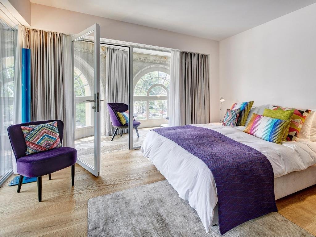 Foto do Hotel Anker Luzern, para representar o post de hotéis em Lucerna. A cama box de casal está no canto direito da foto. Na sua frente há uma poltrona roxa com almofada. Do seu lado esquerdo, há uma mesa de cabeceira com abajur em cima. No fundo também existe uma porta de vidro que dá acesso à outra parte do quarto com várias janelas para a rua, e há mais uma poltrona nesse espaço.