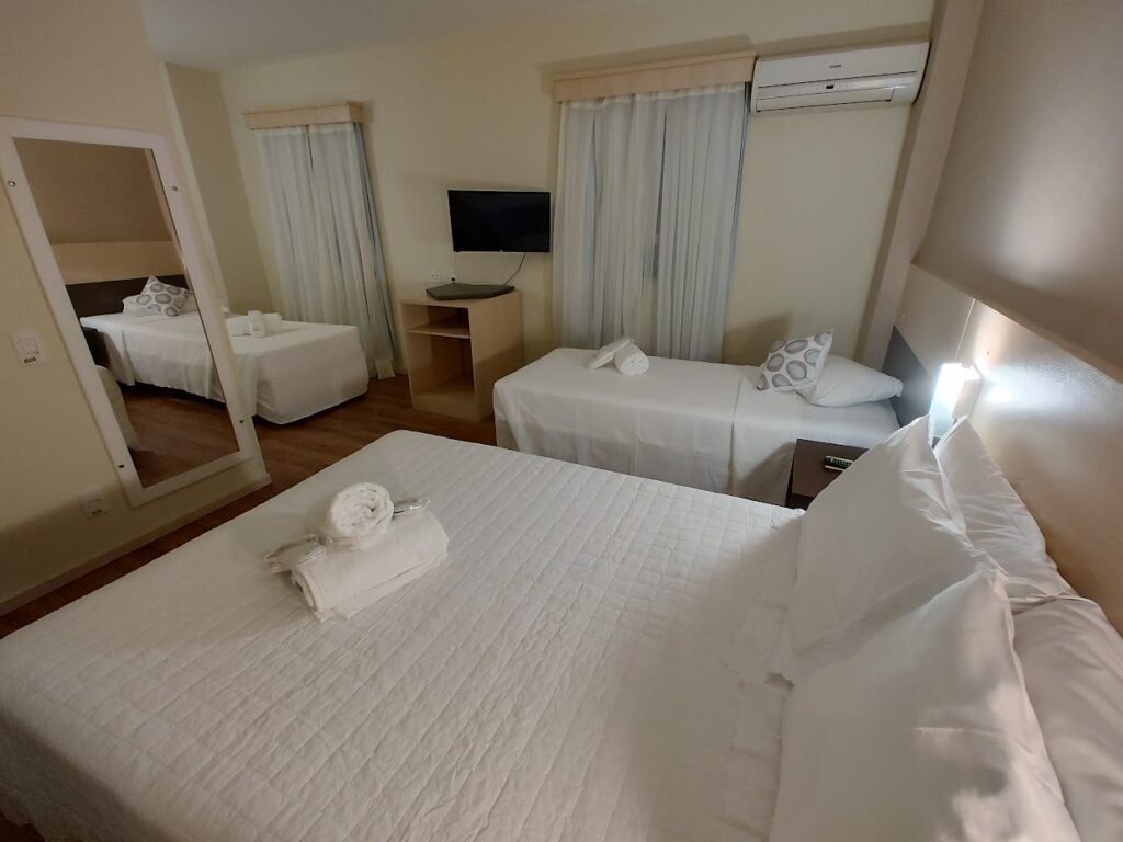 quarto do Bristol Sabrina de Charme Joinville mostrando uma cama de casal no canto inferior direito, ao lado de uma cama de solteiro que está em frente a outra cama de solteiro.