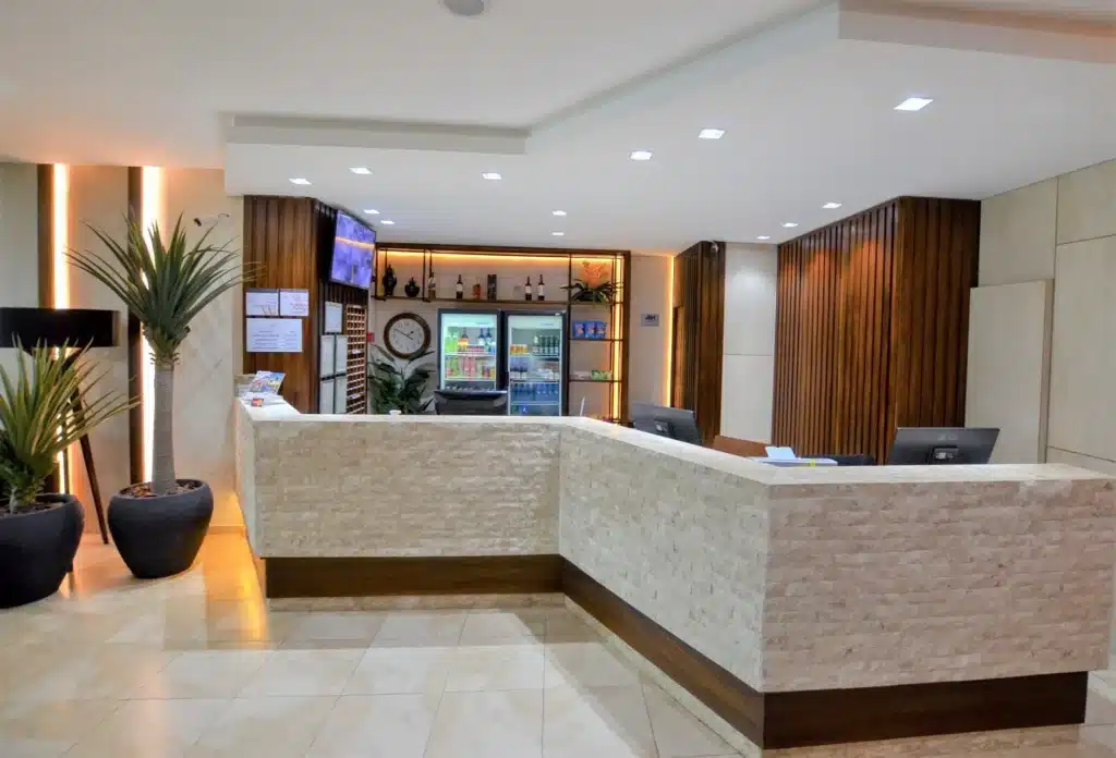 Recepção do hotel com uma bancada, computadores, televisão e máquinas de refrigerante. É possível ver também vasos de plantas. Foto para ilustrar post sobre hotéis em Campo Grande.