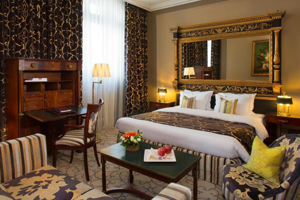 Foto de um quarto do Hotel de la Cignone, para ilustrar o post de hotéis em Genebra. Uma cama king-size está no centro, na direita do quarto. Há um grande espelho retangular na parede atrás. Há uma mesa de cabeceira nos dois lados da cama, com um abajur em cima. Do lado esquerdo, está a janela, e uma escrivaninha de madeira com cadeira e abajur de chão. Na frente da cama há uma pequena mesa de centro com um vaso de flor em cima e prato com duas maçãs. No canto inferior direito, há uma poltrona, do lado da mesa. Outro sofá está de frente para a mesa, no canto inferior esquerdo.