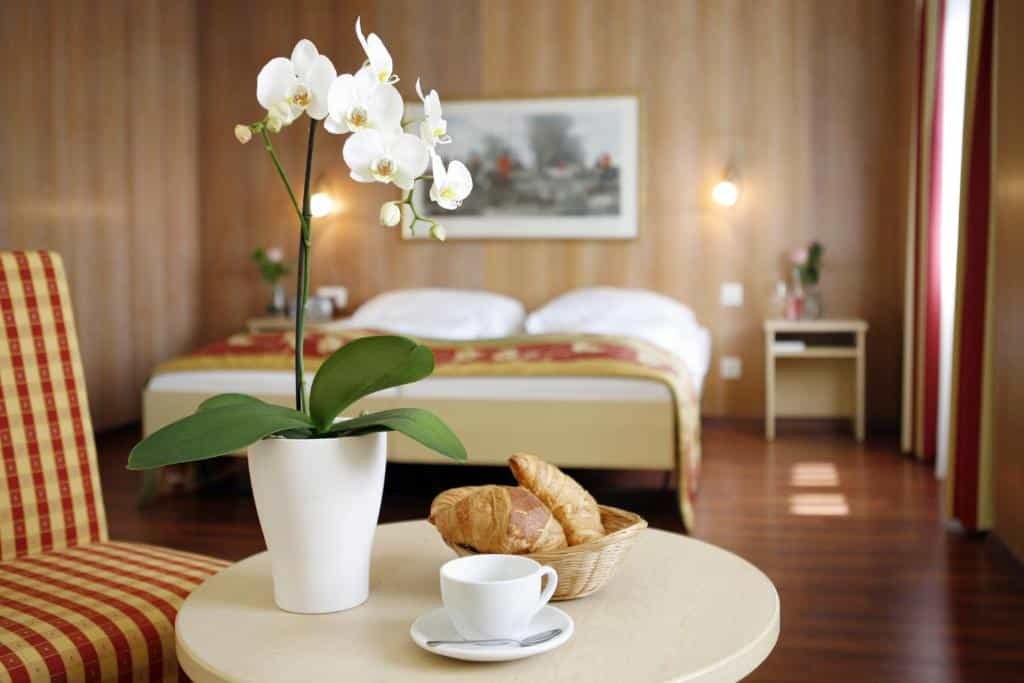Foto do Hotel De la Paix, para representar o post de hotéis em Lucerna. Em foco temos uma mesa pequena redonda com um vaso de flor, xícara e uma cesta com croiassants em sua superfície. No canto da foto, ao seu lado, é possível identificar uma poltrona xadrez. No fundo da foto está a cama e uma escrivaninha em seus dois lados. Uma janela está à sua direita.