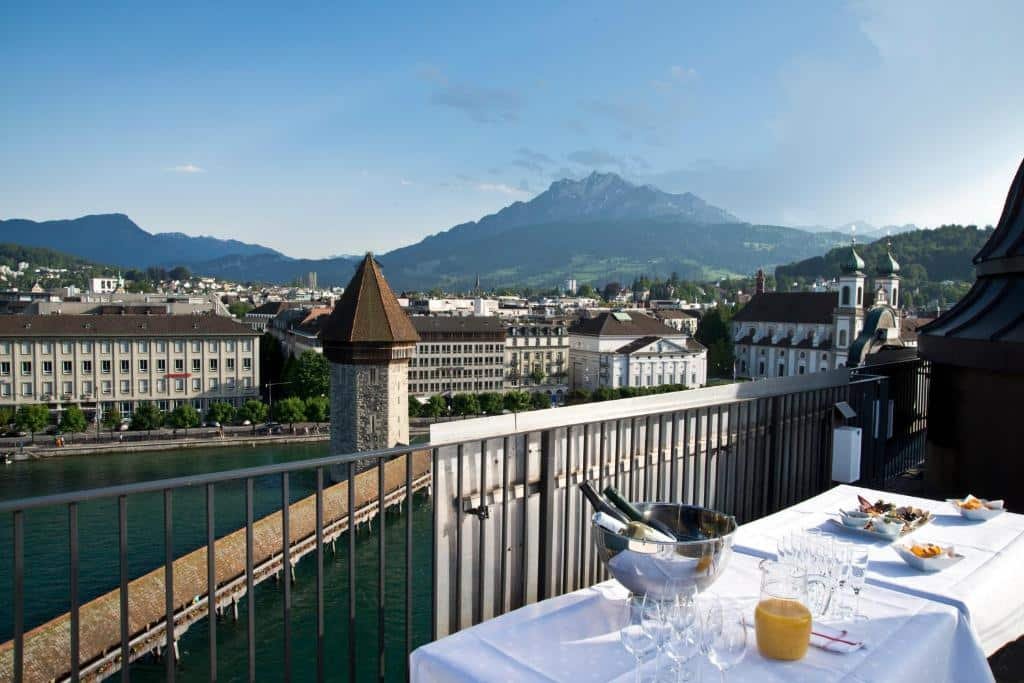 Imagem na varanda do Hotel Des Alpes. Na direita, vemos uma mesa com toalha branca e copos, taças, jarra de suco, petiscos e balde de champagne em cima. Na sua frente, está a grade de ferro da varanda. A vista é para um belo lago e diversas casas e prédios da cidade de Lucerna. As montanhas também estão ao fundo.