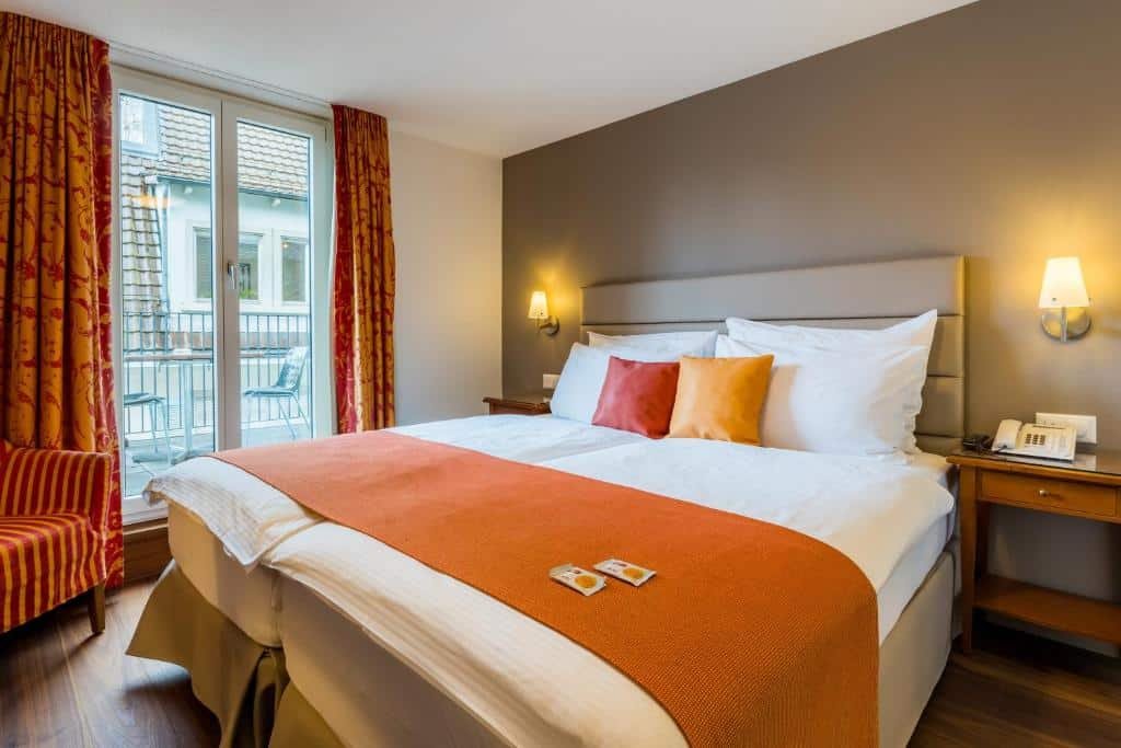 Foto do Hotel Des Alpes, para representar o post de hotéis em Lucerna. A cama é de casal e está no centro da foto, encostada na parede à direita. Há uma mesa de cabeceira acompanhada de uma luminária em seus dois lados. A janela com varanda está à esquerda da cama No canto inferior esquerdo da foto ainda há uma poltrona.