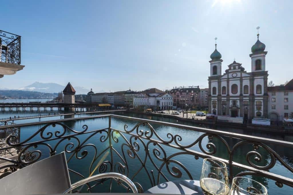 Vista para o rio em uma varanda no Hotel des Balances, em Lucerna. Está e manhã, o sol raia em direção da foto. Há uma mesa e duas cadeiras na varanda, depois o rio e construções tradicionais do outro lado.
