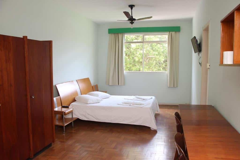 Quarto do Hotel do CEE. No lado esquerdo há um armário de madeira, uma cama de casal e ao fundo uma janela. No lado direito há uma mesa, duas cadeiras, uma TV e a porta para o banheiro. Imagem para ilustrar o post hotéis em Viçosa.