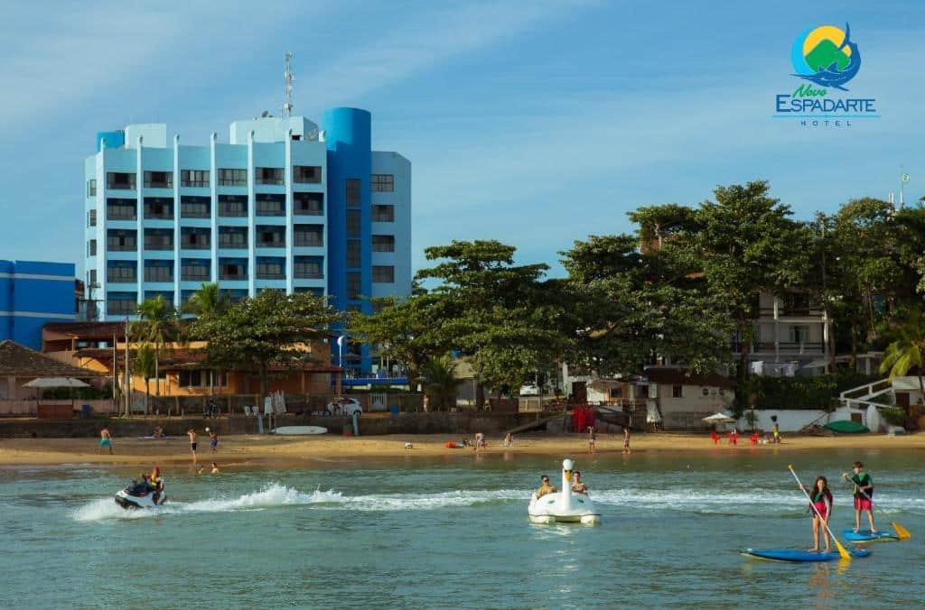 Frente do Hotel Espadarte. O mar na frente com pessoas andando de pedalinho, jet ski e stand up paddle. No fundo pessoas na areia e um quiosque, atrás árvores uma rua e o hotel. Foto para ilustrar post de pousadas em Iriri (ES).