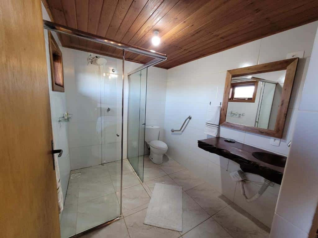 Banheiro do Hotel Fazenda Boutique Terra do Gelo com chuveiro de vidro, vaso sanitário com barras de apoio e bancada de pia de madeira. Paredes de azulejos brancos e teto de madeira são visíveis.