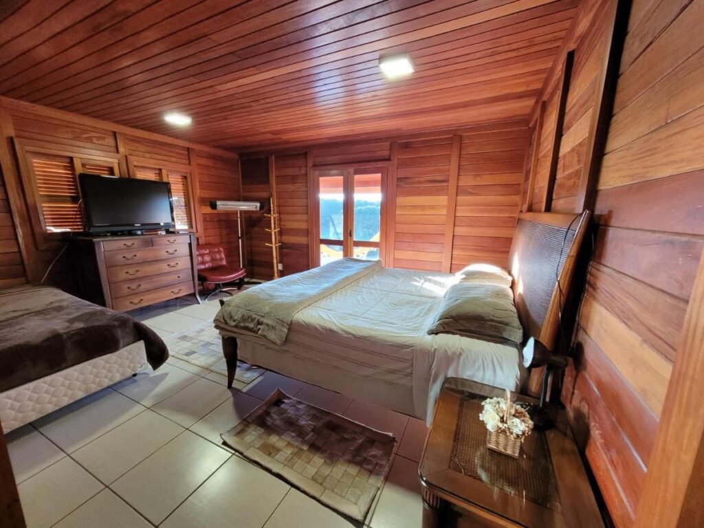 Um aconchegante quarto de madeira no Hotel Fazenda Boutique Terra do Gelo com uma cama grande, uma cama pequena, uma TV em uma cômoda e uma janela com vista. Um tapete e uma mesa de cabeceira adornam o quarto.