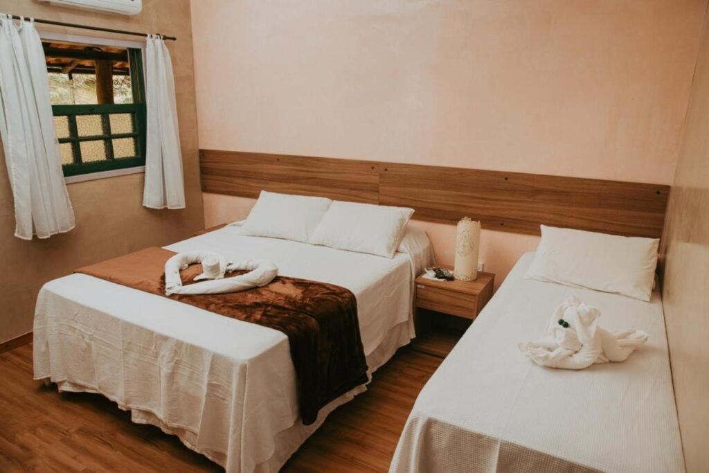 A imagem mostra um quarto aconchegante do Hotel Fazenda Cheiro Verde com uma cama de casal e uma cama de solteiro, ambas cobertas com roupas de cama brancas. Sobre a cama de casal, há uma manta marrom e uma toalha dobrada em formato de cisne. A cama de solteiro também possui uma toalha dobrada em formato decorativo. Entre as camas, há uma mesa de cabeceira de madeira com uma luminária de design simples. O quarto tem paredes em tons suaves de bege e rosa, com piso de madeira clara, criando um ambiente acolhedor e rústico. Uma janela com cortinas brancas permite a entrada de luz natural.