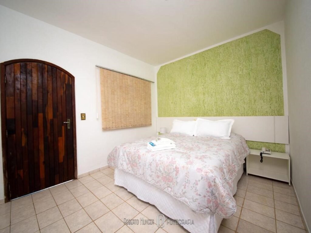 A imagem mostra um quarto simples e acolhedor do Hotel Fazenda Colina, com uma cama de casal ao centro coberta por uma colcha floral em tons claros. A parede atrás da cama é decorada com uma textura em verde suave, contrastando com as paredes brancas ao redor. Ao lado da cama, há uma pequena mesa de cabeceira branca. A porta de entrada é de madeira escura com um formato arredondado no topo, criando um toque rústico. O chão é de cerâmica clara, e há uma janela com uma persiana fechada ao lado da cama.