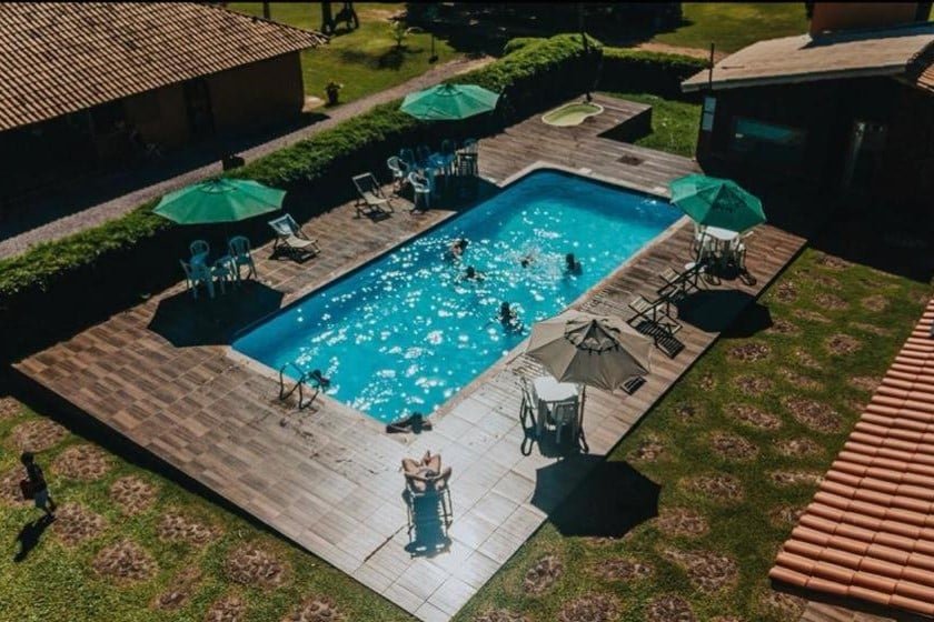 A imagem mostra uma piscina retangular ao ar livre em um dia ensolarado, cercada por um amplo deque de piso de pedra. Várias pessoas estão dentro da piscina aproveitando o momento, enquanto ao redor há espreguiçadeiras e mesas com guarda-sóis verdes para quem deseja descansar à sombra. A piscina é cercada por uma área verde bem cuidada, e ao fundo, há construções com telhados de telha vermelha. O ambiente é tranquilo, ideal para lazer e relaxamento, com uma atmosfera acolhedora de campo ou fazenda. Esse é o Hotel Fazenda Coninho, um dos hotéis-fazenda em Minas Gerais.