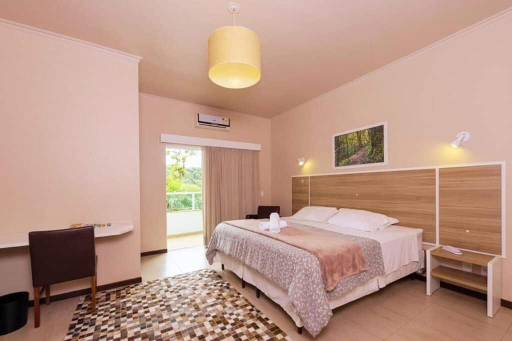 Um quarto do Hotel Fazenda Dona Francisca com uma cama, escrivaninha, cadeira, mesas de cabeceira, um tapete e um ar condicionado de parede. Uma porta se abre para uma sacada com vegetação.