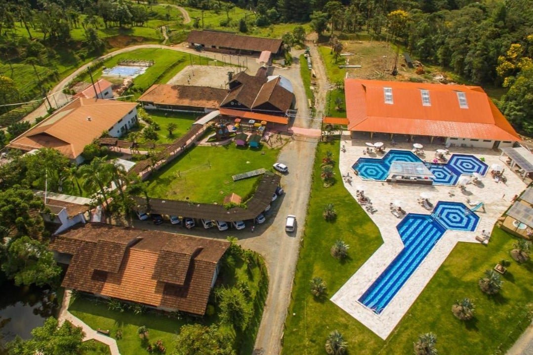 Vista aérea do Hotel Fazenda Dona Francisca com piscinas, edifícios com telhados vermelhos, área verde e área de estacionamento, cercado por vegetação e árvores. Esse é um dos hotéis fazenda em Santa Catarina.