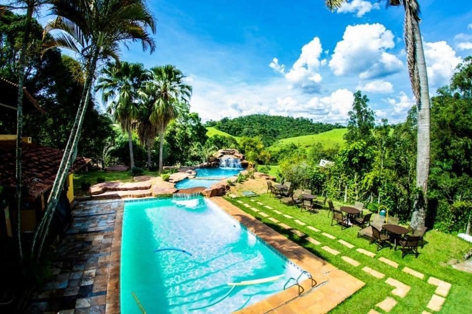 A imagem mostra uma área externa com duas piscinas cercadas por natureza exuberante. A piscina menor, em primeiro plano, está localizada ao lado de um caminho de pedras e cercada por mesas e cadeiras dispostas sobre um gramado bem cuidado. Ao fundo, uma piscina maior tem formato orgânico e inclui uma pequena cachoeira artificial decorada com pedras. Palmeiras altas se erguem ao redor das piscinas, e colinas verdes compõem o cenário ao longe, com um céu azul e nuvens brancas que complementam a paisagem tropical e relaxante. Esse é o Hotel Fazenda Horizonte Belo, um dos hotéis-fazenda em Minas Gerais.