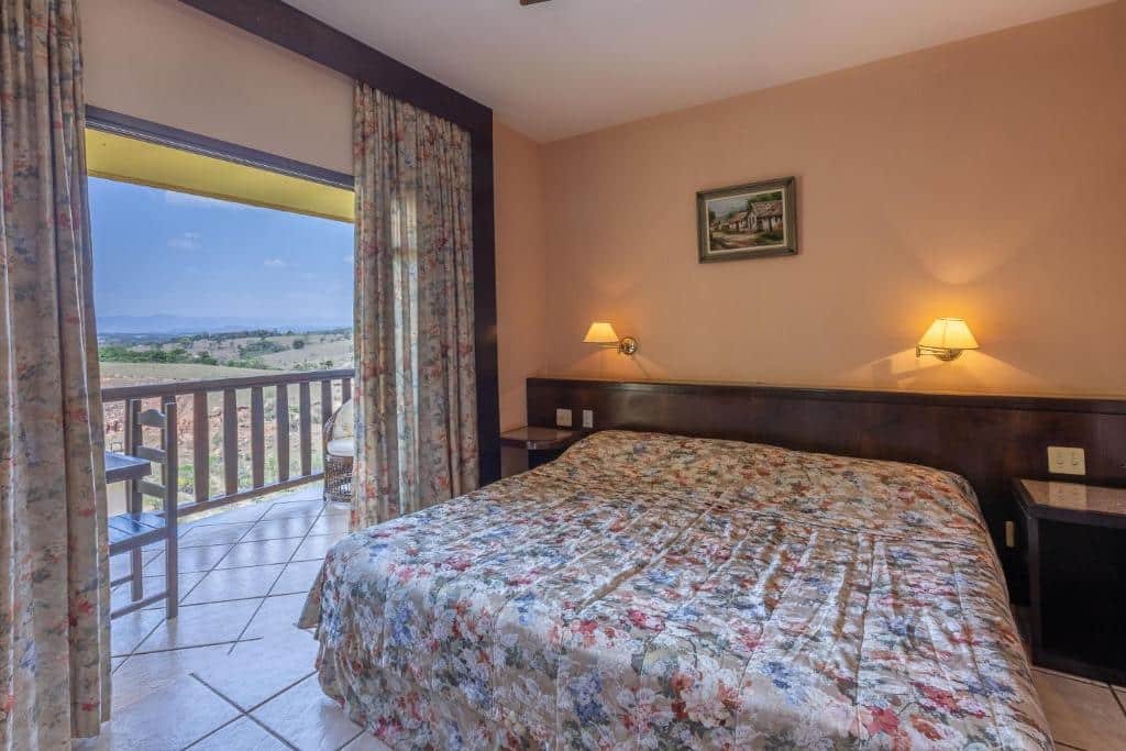A imagem mostra um quarto com uma cama de casal coberta por uma colcha floral em tons suaves no Hotel Fazenda Horizonte Belo. Ao lado da cama, há uma grande porta de vidro que leva a uma varanda com vista para uma paisagem de montanhas e campos. Cortinas florais enquadram a porta, complementando a decoração do quarto. Duas luminárias de parede, uma de cada lado da cama, oferecem iluminação suave e aconchegante. Na parede acima da cama, há um pequeno quadro decorativo. O chão é de azulejo claro, e a varanda está equipada com uma cadeira, proporcionando um espaço para relaxar e apreciar a vista.