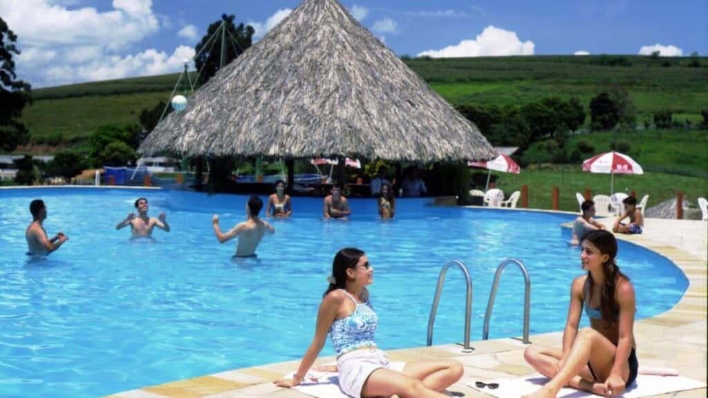 A imagem mostra uma grande piscina ao ar livre em um dia ensolarado no Hotel Fazenda Menino da Porteira. Duas mulheres estão sentadas na borda da piscina, conversando e tomando sol. Ao fundo, algumas pessoas estão dentro da água, jogando e se divertindo. No centro da piscina, há um bar molhado coberto por um teto de palha, oferecendo sombra para os banhistas. Ao redor, guarda-sóis brancos e vermelhos cobrem mesas com cadeiras. A paisagem ao fundo inclui colinas verdes, criando um cenário relaxante e natural.