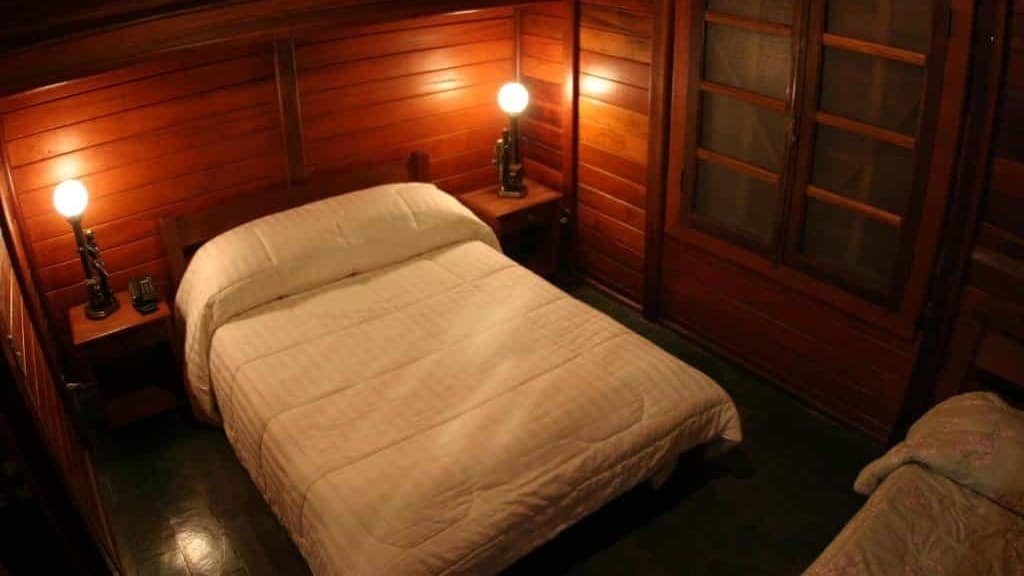 A imagem mostra um quarto rústico com paredes de madeira no Hotel Fazenda Menino da Porteira. No centro, há uma cama de casal coberta com um edredom branco. Ao lado da cama, dois criados-mudos com luminárias de abajur clássico emitem uma luz suave, criando um ambiente aconchegante. As janelas, também de madeira, possuem vidro, e ao lado direito, parcialmente visível, há outra cama coberta com uma colcha clara. O ambiente é acolhedor, com um estilo simples e confortável, ideal para descanso em um espaço mais intimista.