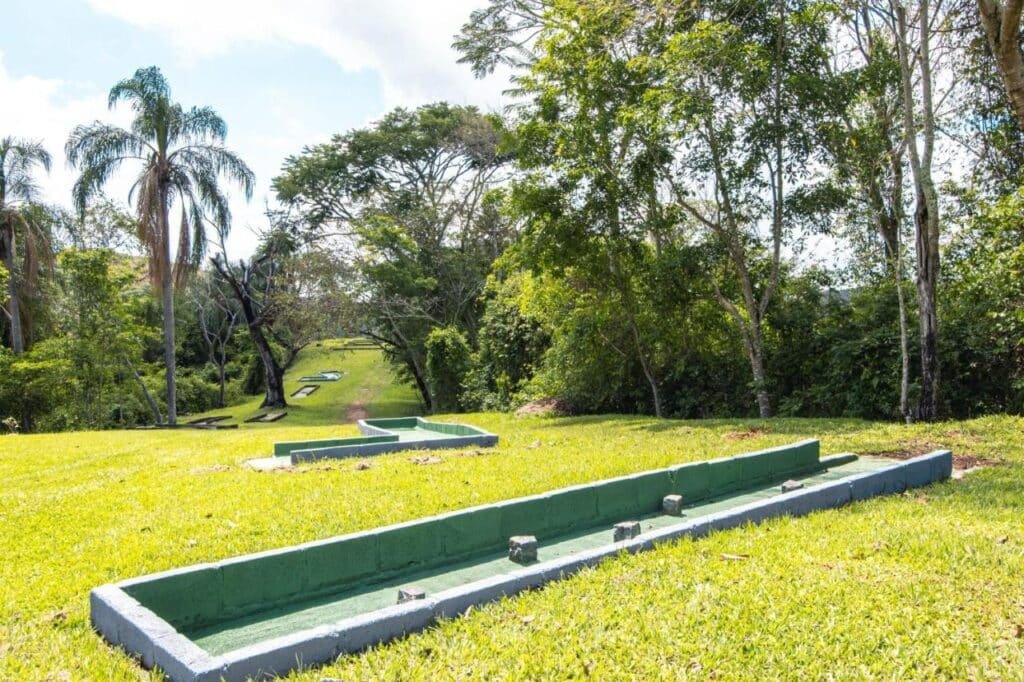 A imagem mostra um campo gramado ao ar livre com estruturas retangulares baixas e pintadas de verde, que parecem ser pistas de minigolfe. As pistas estão dispostas em diferentes níveis, acompanhando a inclinação suave do terreno. Ao fundo, há árvores grandes e palmeiras, criando um ambiente natural e tranquilo. O céu está claro e azul, e a área ao redor das pistas é cercada por vegetação densa, tornando o espaço do Hotel Fazenda Minas Realconvidativo para atividades recreativas ao ar livre.