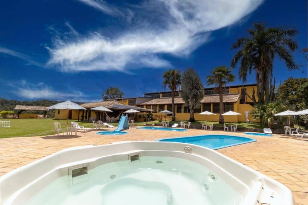 A imagem mostra uma área de lazer externa com uma banheira de hidromassagem em primeiro plano e uma piscina ao fundo. Ao lado da piscina, há um pequeno escorregador azul para crianças. A área ao redor da piscina está cercada por espreguiçadeiras brancas com guarda-sóis, criando um ambiente convidativo para relaxar. Ao fundo, há uma grande casa de estilo rústico, pintada de amarelo, com telhado de telhas de barro, cercada por palmeiras e vegetação verde. O céu está claro, com algumas nuvens, sugerindo um dia ensolarado e agradável. Esse é o Hotel Fazenda Minas Real, um dos hotéis-fazenda em Minas Gerais.